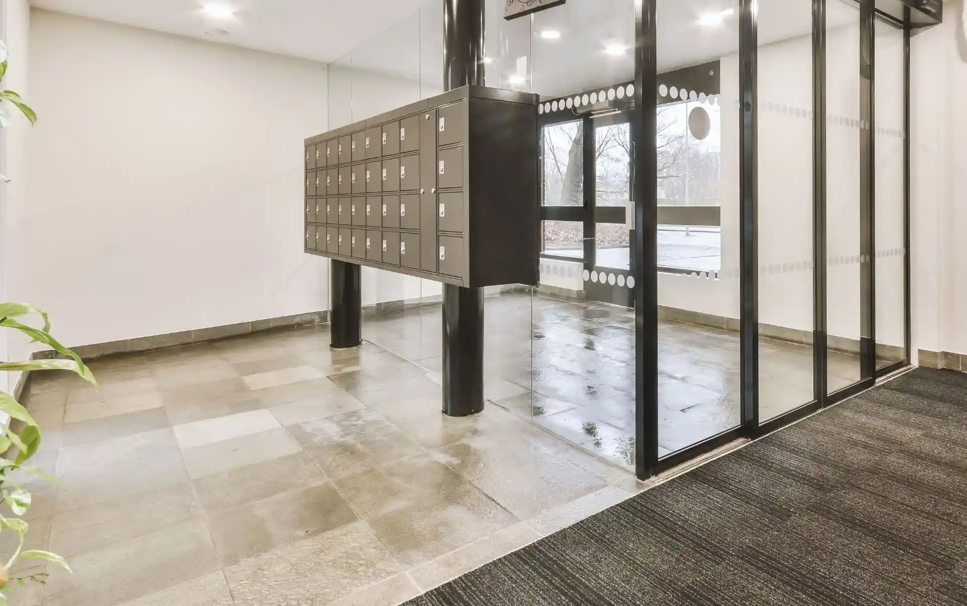 Hausmeisterservice-Reutlingen-Stuttgart-STR24-Facility-Management Moderner Eingangsbereich eines Gebäudes mit einer Wand aus Postfächern und großen Glasfenstern. Der Boden ist gefliest und wirkt sauber und gepflegt. Die Glaswände lassen viel Licht in den Raum und schaffen eine helle und einladende Atmosphäre. Dieses Bild repräsentiert den professionellen Hausmeisterservice der von STR24 Gebäudemanagement aus Stuttgart angeboten wird. Wir kümmern uns um die Wartung und Sauberkeit von Ihren Immobilien.