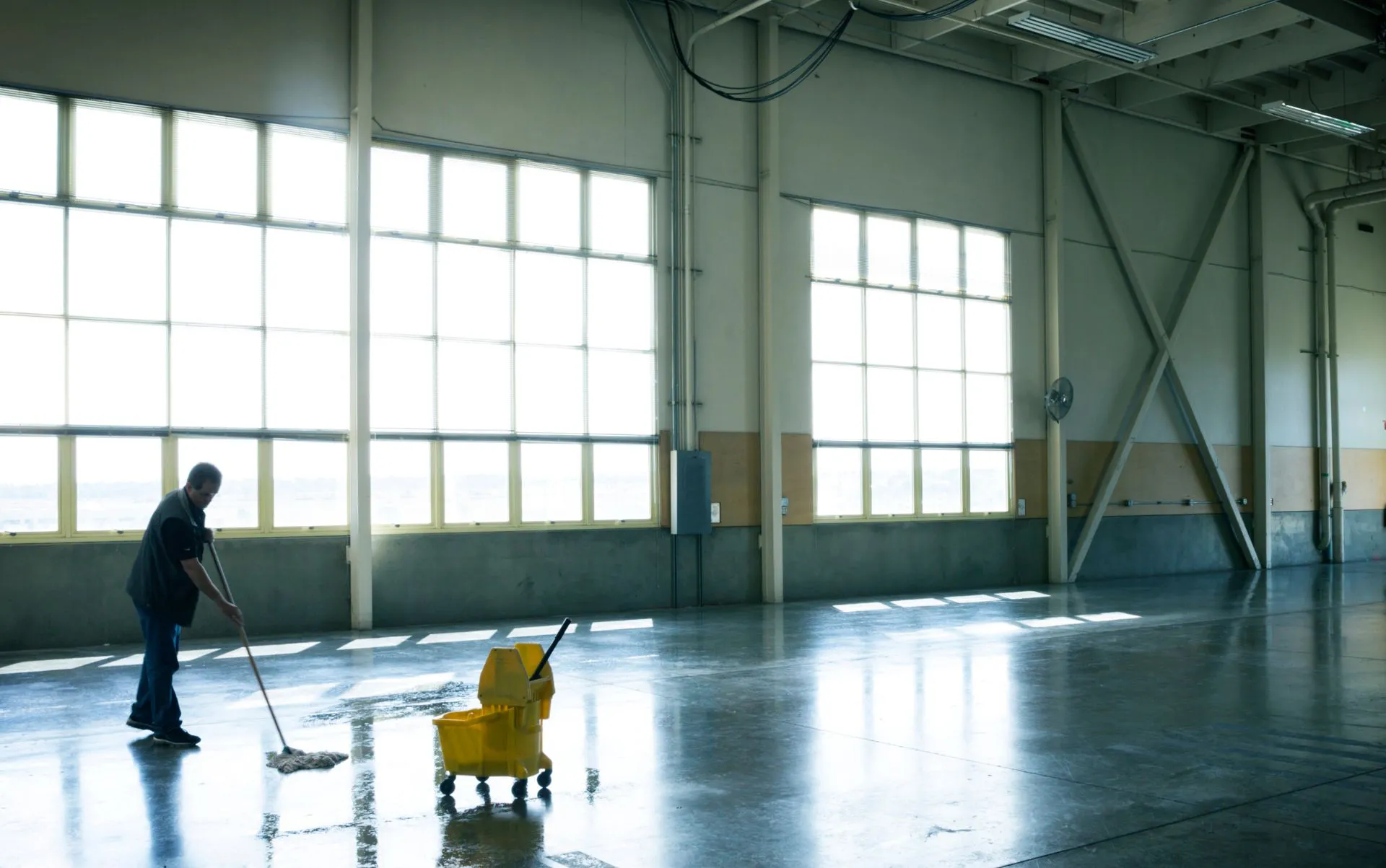 Professionelle Reinigungskraft von STR24 Gebäudemanagement bei der Industriereinigung, reinigt mit einem Mopp und einem gelben Putzwagen einen großen, sonnendurchfluteten Industriehallenboden.